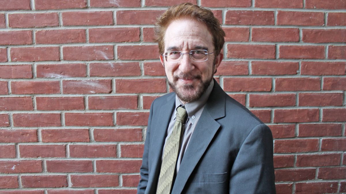 Man in a suit and glasses stands in front of a brick wall