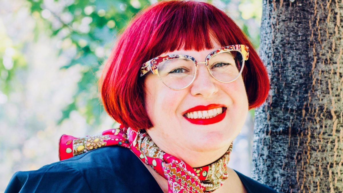 A smiling woman with red hair and distinctive glasses.