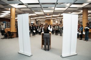 Flanked by pop-up banners, a woman stands at a podium, addressing a large crowd.