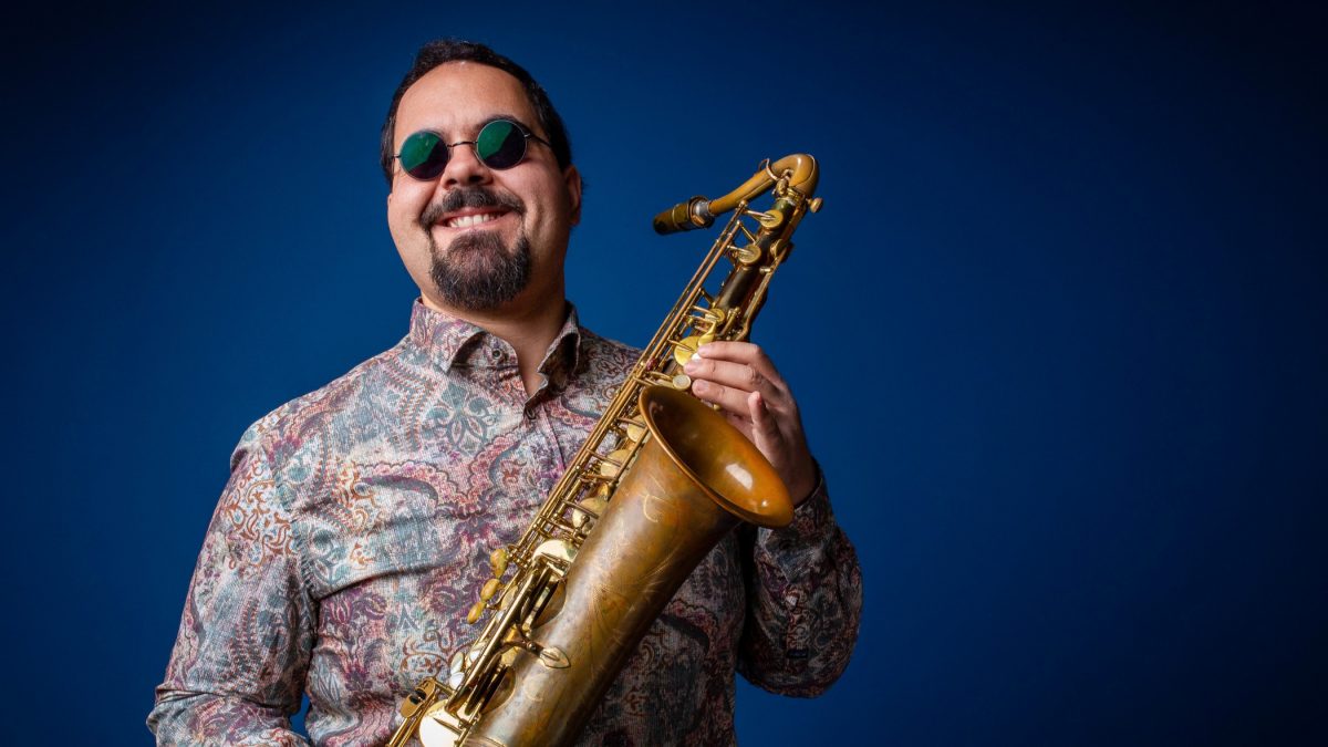 A man smiles while wearing sunglasses and holding a saxophone