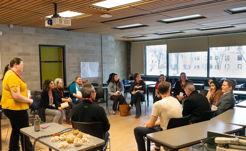 A group of students sit in chairs in a circle while one stands at the front.