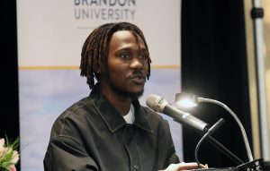 A young man speaks at a microphone. A banner behind him says "Brandon University Alumni"