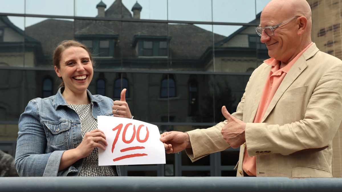 Two people give thumbs up while holding a piece of paper on which is written a large red 100.