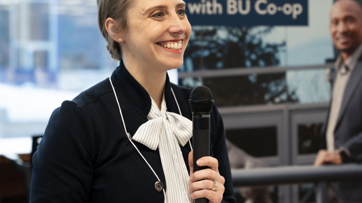 A woman smiles while holding a microphone, in front of a banner that promotes the BU Co-op program.