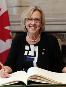 A woman sits at a desk and smiles as she writes in a book. A Canadian flag can be seen behind her on the left of the picture.
