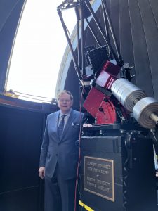 A man in a suit stands next to a large telescope. Behind him there is an opening in a large protective dome.