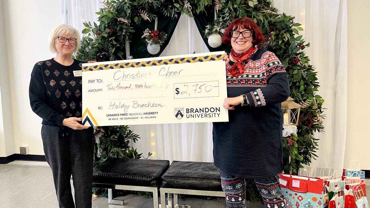 Two women stand in front of holiday garland with an oversized promotional cheque that reads "$2,750" made out to Christmas Cheer