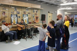 A family watches a fiddler and a guitarist perform in front of the Sports Wall of Fame in the Healthy Living Centre