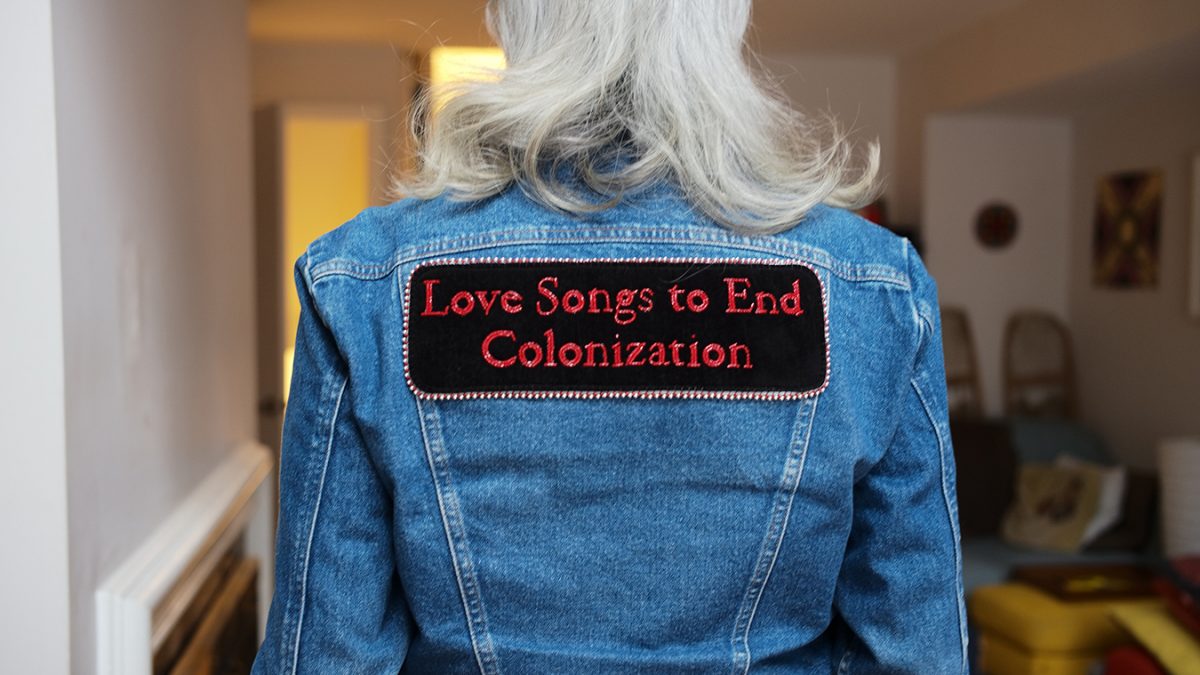 The back of a person with grey hair, wearing a denim jacket with a patch that says "Love Songs to End Colonization"