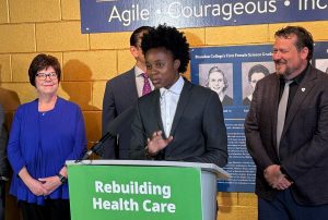 A person speaks while standing at a lectern with three people behind them