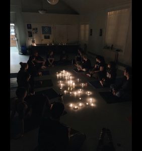 A group of people sit in a circle in a dark room, with candles on the floor in the middle