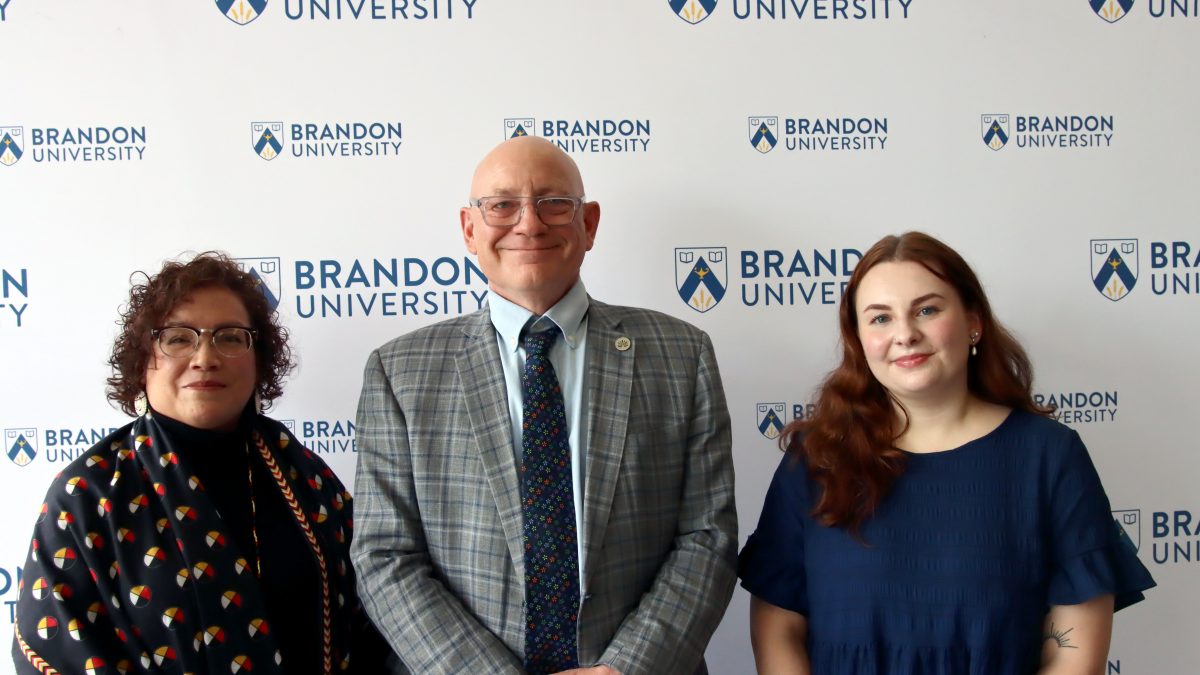 Three people stand in front of a Brandon University backdrop
