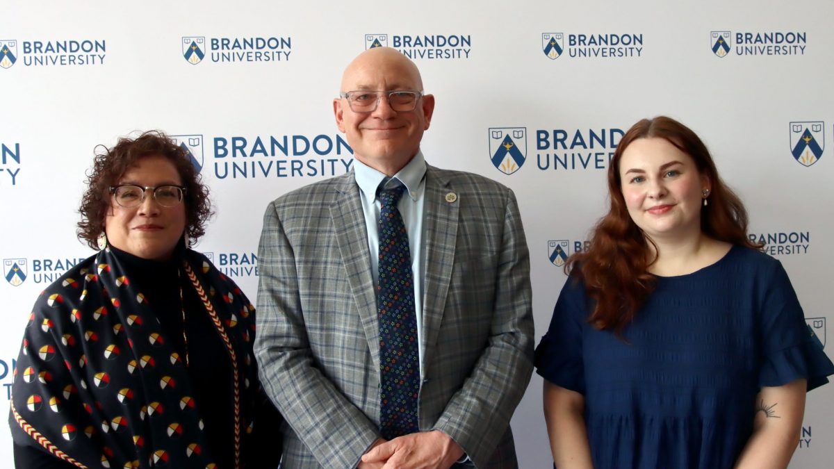 Three people stand in front of a Brandon University backdrop