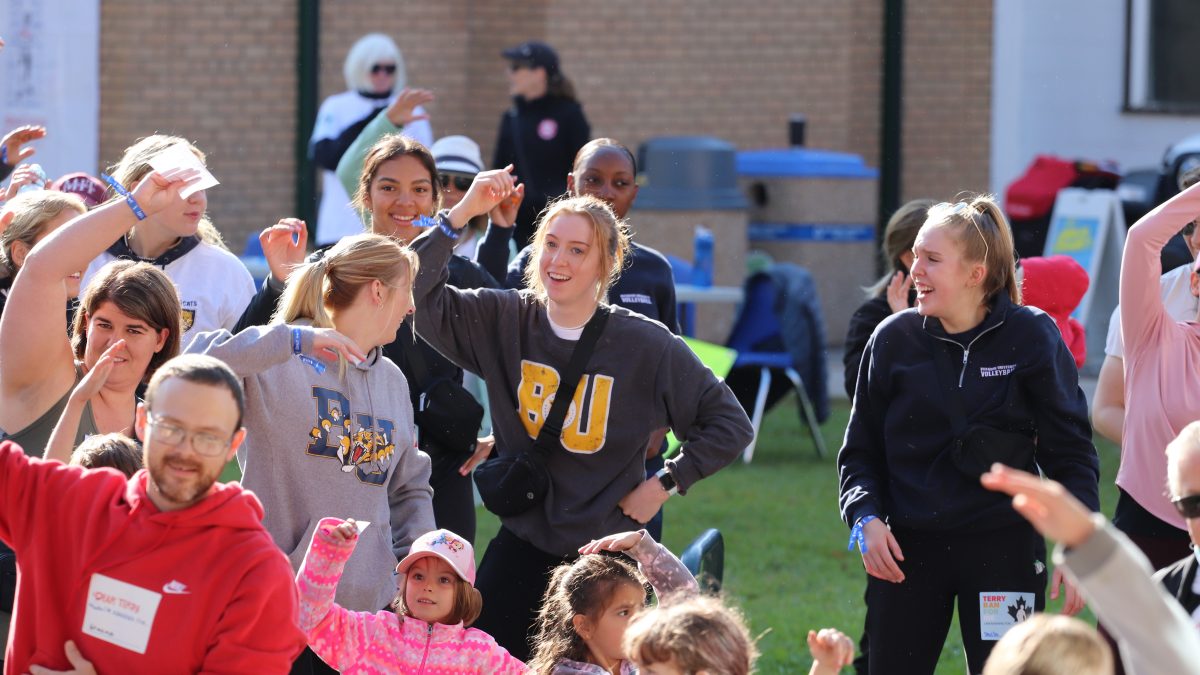 A large number of people stretch outdoors. A woman wearing a BU sweatshirt smiles at the centre if the image