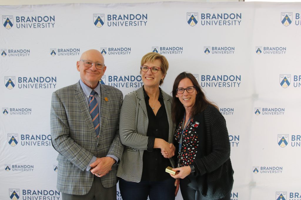 Three people post in front of a backdrop with the Brandon University logo.