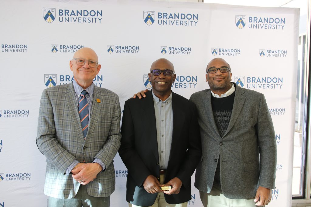 Three people post in front of a backdrop with the Brandon University logo.