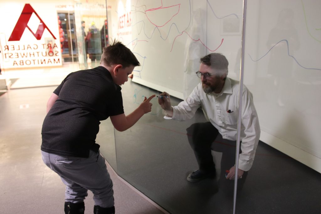 A boy points to a glass wall, while a man at the other side draws with a marker where the boy is pointing