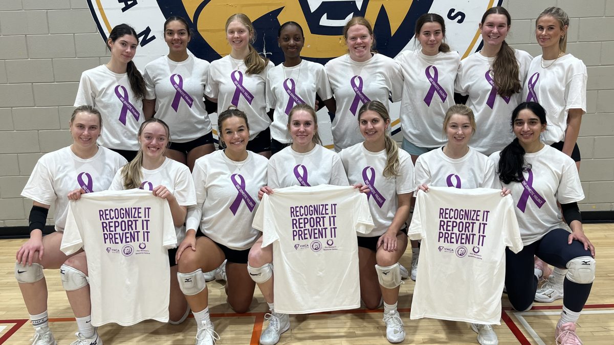 A row of athletes stands in a gym with another row in front of them. They are wearing white t-shirts with an image of a purple ribbon on the front. Three of the athletes are holding shirts so the back can be seen. The shirts say "Recognize It, Report It, Prevent It."