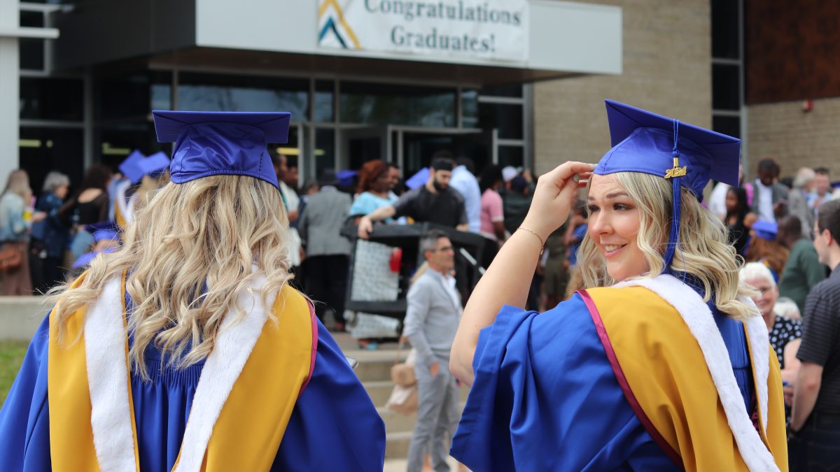 Two female graduates look at each other outside the Healthy Living Centre at BU where a banner hangs, reading Congratulations Graduates.