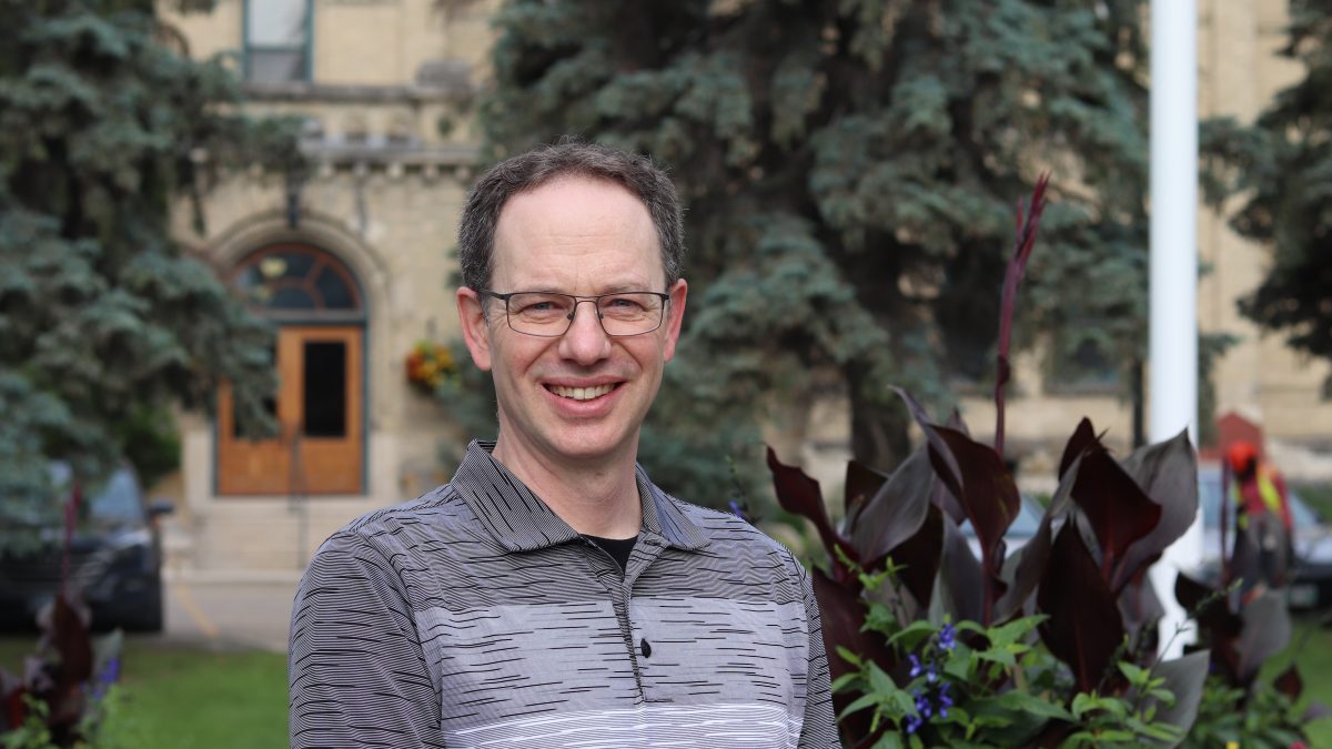 A man smiles while posing in front of plants and a brick building