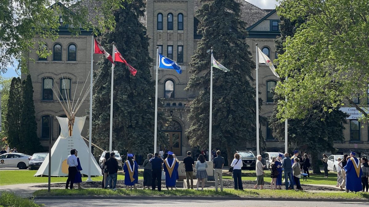 A crowd of people, some of them in graduation robes, gather in front of a large brick building. There are a tipi and several flags in front of the building.
