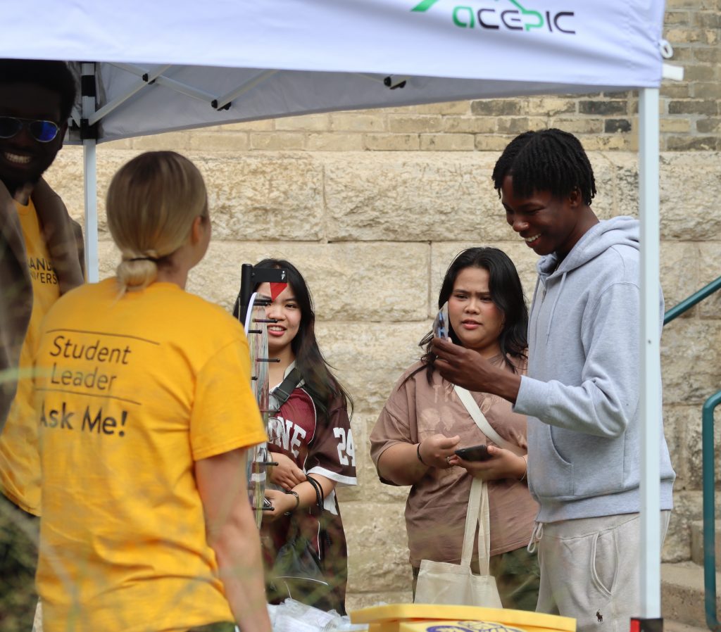 People visit a booth. One of the people in the booth wears a t-shirt saying "Student Leader. Ask me!"