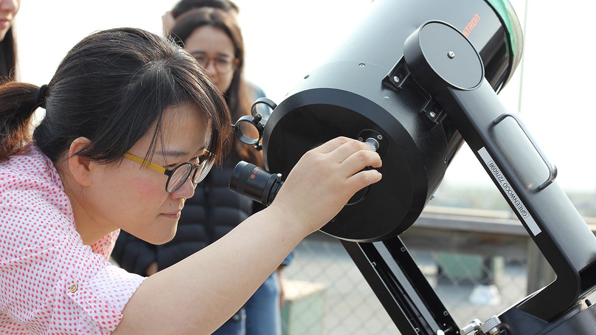 A woman leans forward to look in the eyepiece of a telescope