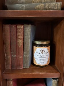 A jar of honey, labelled Bee U, sits on a shelf next to vintage hardcover books.