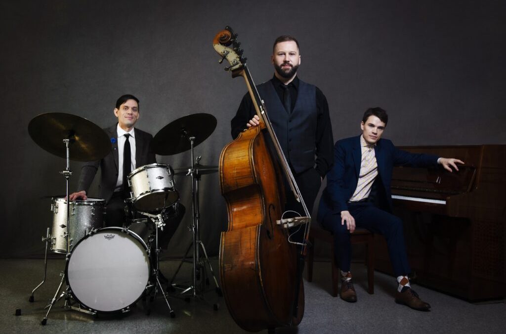 A man sits at a drums et, a man stands with an upright bass, and another man sits at a piano
