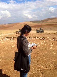 A woman is seen from the back writing in a notebook in the desert. Armoured vehicles are in the distance.