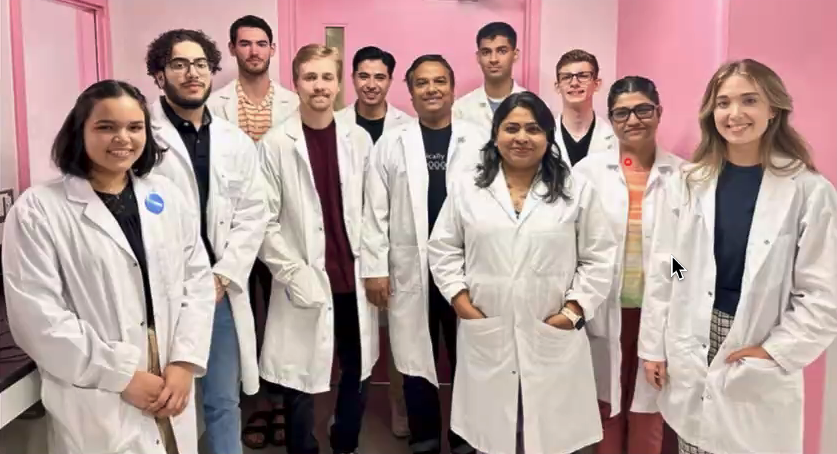 A group of people pose in lab coats in a pink painted room