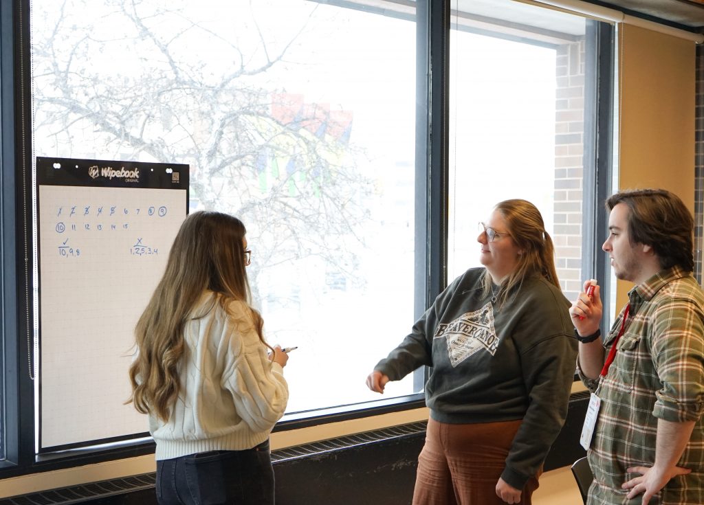 Two students discuss with a third, who is in front of a flip chart.