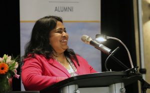 A woman speaks at a microphone. A banner behind her says "Brandon University Alumni"