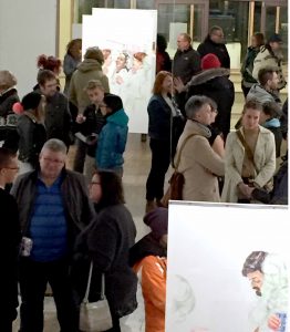 A crowed of people stand in the art gallery, with images on display in the foreground and in the back of the photo