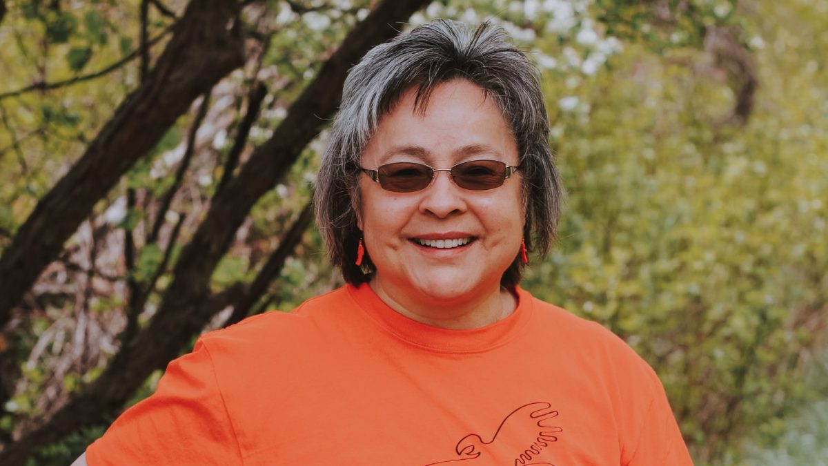 A woman standing in front of trees smiles while wearing an orange shirt
