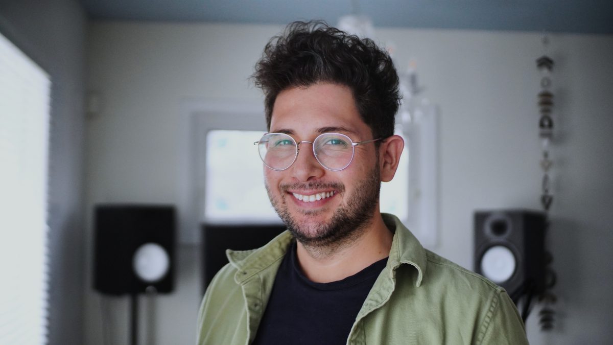 A man smiles in a white room. Audio speakers can be seen behind him