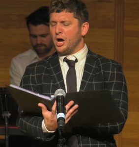 A singer performs into a microphone as he reads the music book in front of him. Another performer is seen in the background.