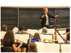 A woman stands in front of a classroom of musicians with a conductor's baton.