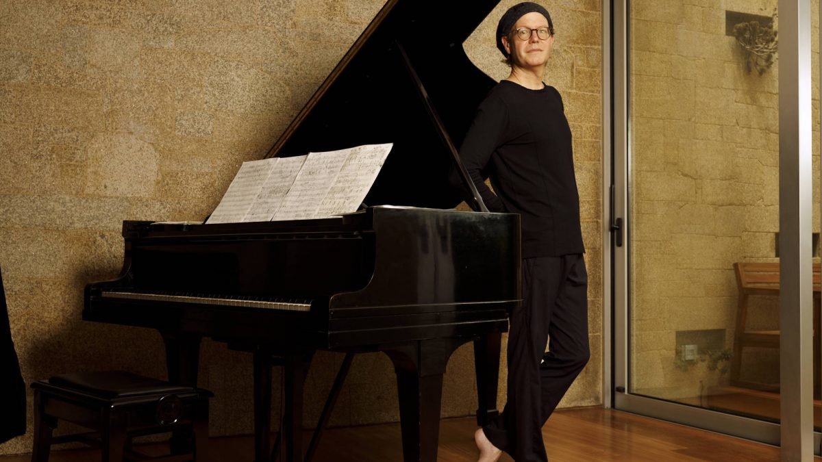 A man, dressed in black and in bare feet, stands next to a piano