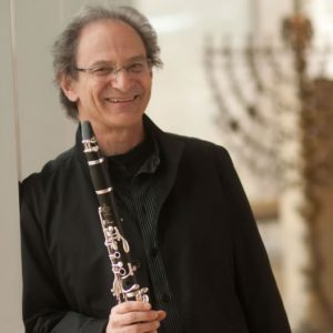 A man leans against the wall and smiles while holding a clarinet in one hand