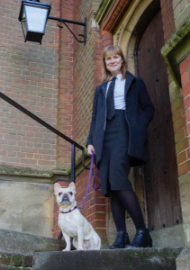 A woman stands in the doorway of a brick building with a dog sitting beside her