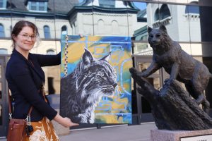 A woman holds up a painting of a Bobcat, while standing next to a statue of a Bobcat.