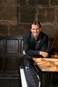 Murray McLachlan leans on a piano and smiles while standing in front of a stone and wooden wall.